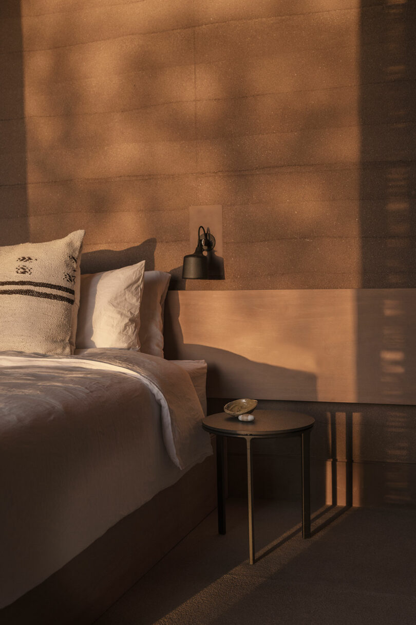A warmly lit bedroom with a double bed, white bedding, a patterned pillow, and a small black side table holding a decorative bowl and object; shadows are cast on the tan wall.