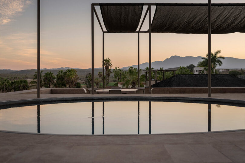 Circular pool and shaded area with a view of palm trees and distant mountains at sunset.