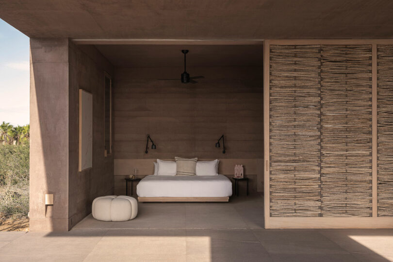 Minimalist bedroom with open-wall design, featuring a bed with white linens, two wall-mounted lamps, a ceiling fan, and a wicker textured partition. The space opens to an outdoor view.