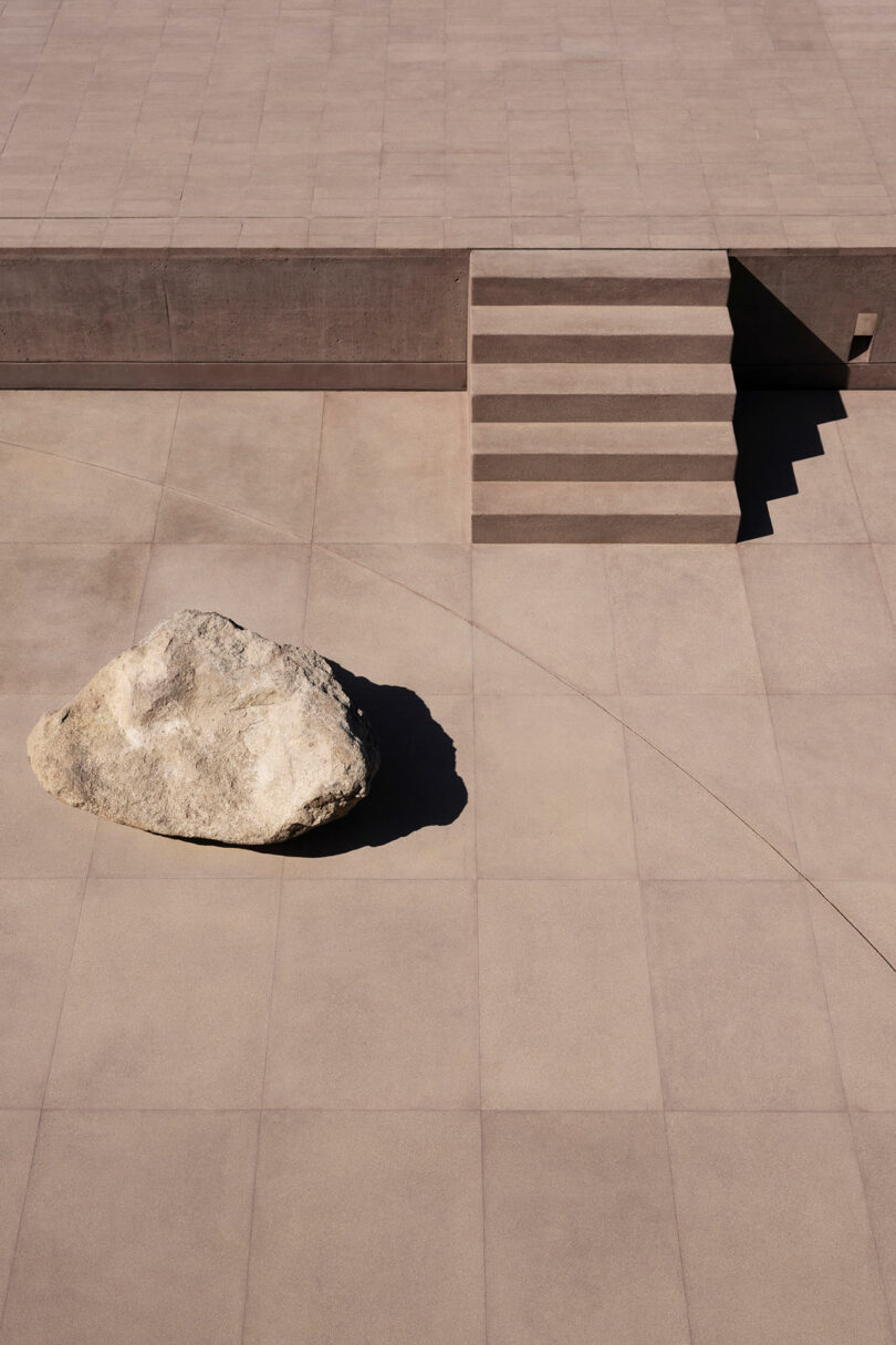 A large rock sits on a minimalist, geometric concrete patio next to a small set of stairs, casting a prominent shadow under daylight.
