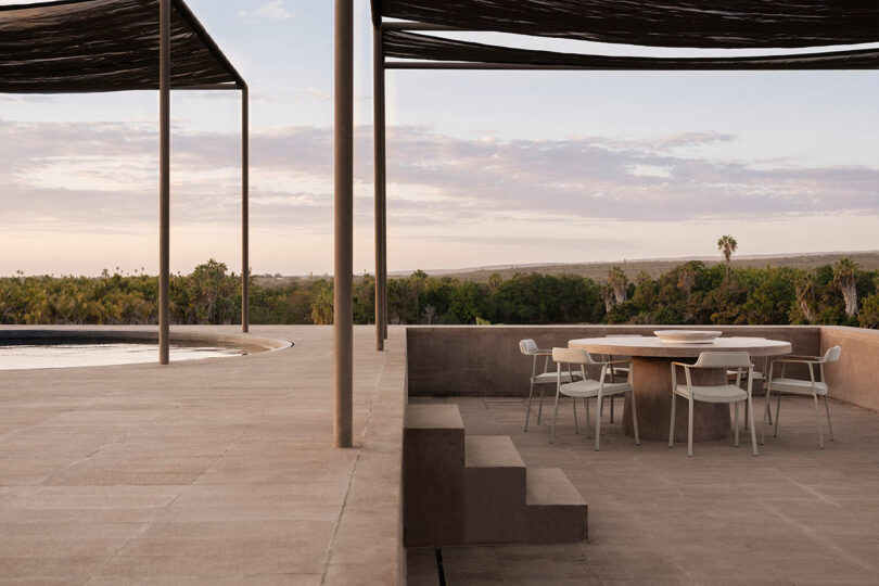 Outdoor patio with a circular stone table surrounded by white chairs. Overhead, slatted pergolas provide shade. The view overlooks greenery and a distant horizon under a cloudy sky.