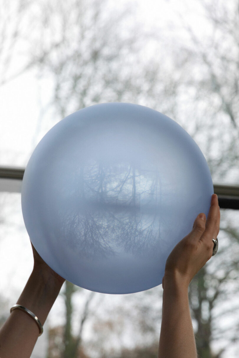 Hands are holding up a translucent blue balloon with faint reflections of trees and sky visible on its surface.
