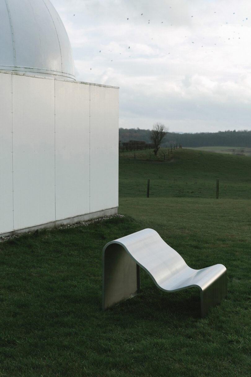 Curved metal bench on a grassy field near a white building with a dome. Overcast sky and distant landscape in the background.
