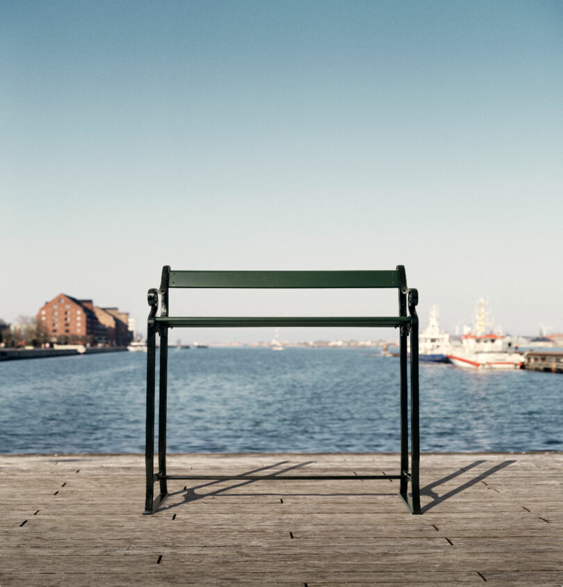 A green, backless bench on a wooden dock overlooks a calm body of water with buildings and ships in the background under a clear sky.