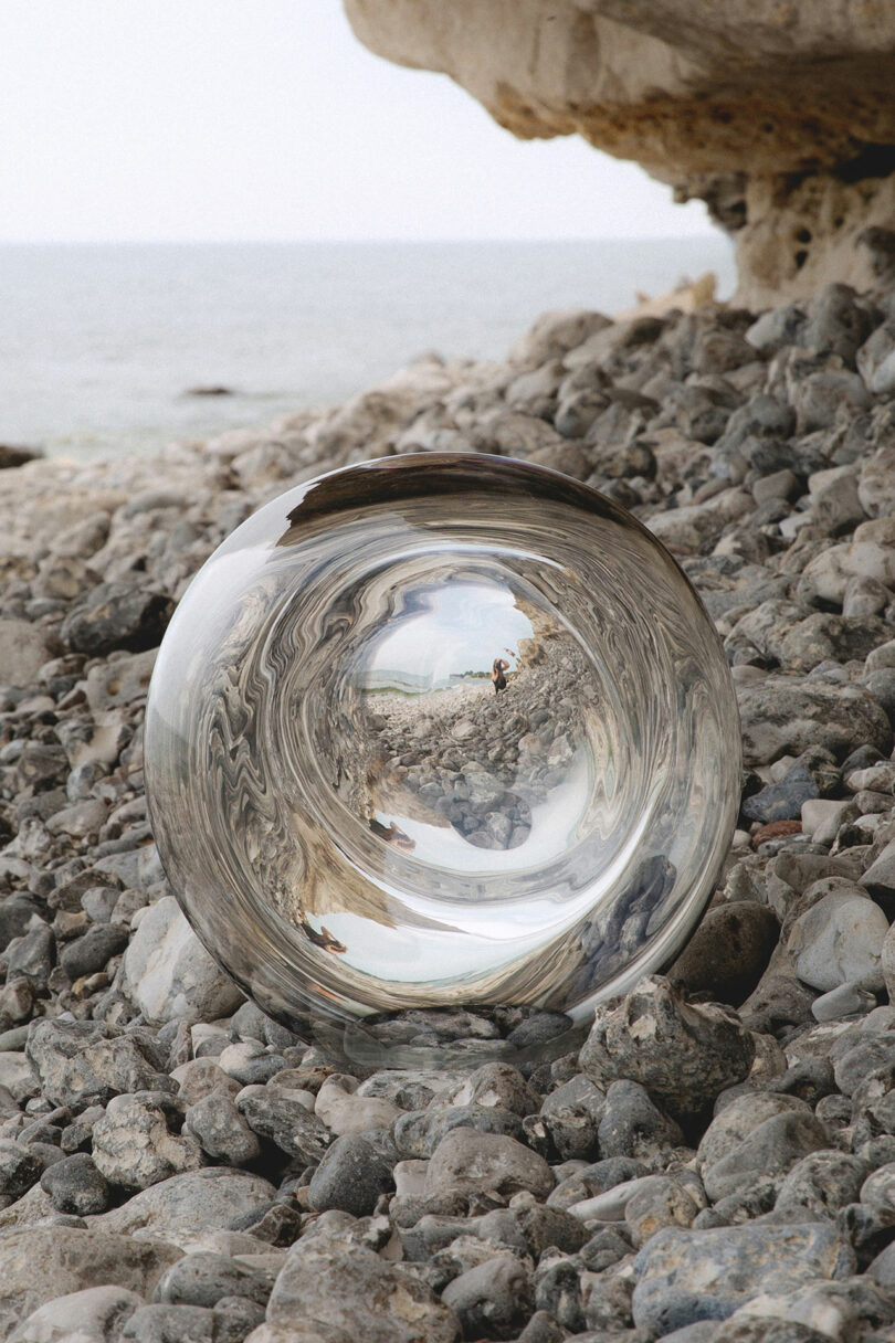 A reflective sphere rests on a rocky beach, surrounded by pebbles, with the sea visible in the background.