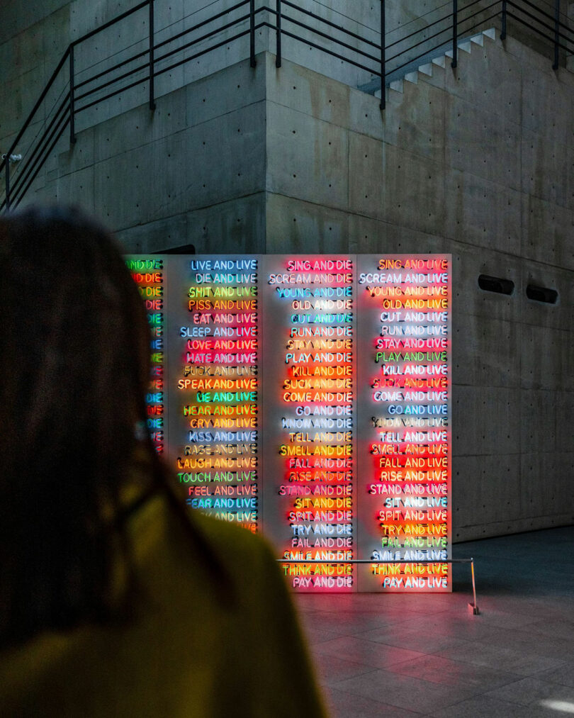 Person viewing a large wall installation with colorful neon text phrases like