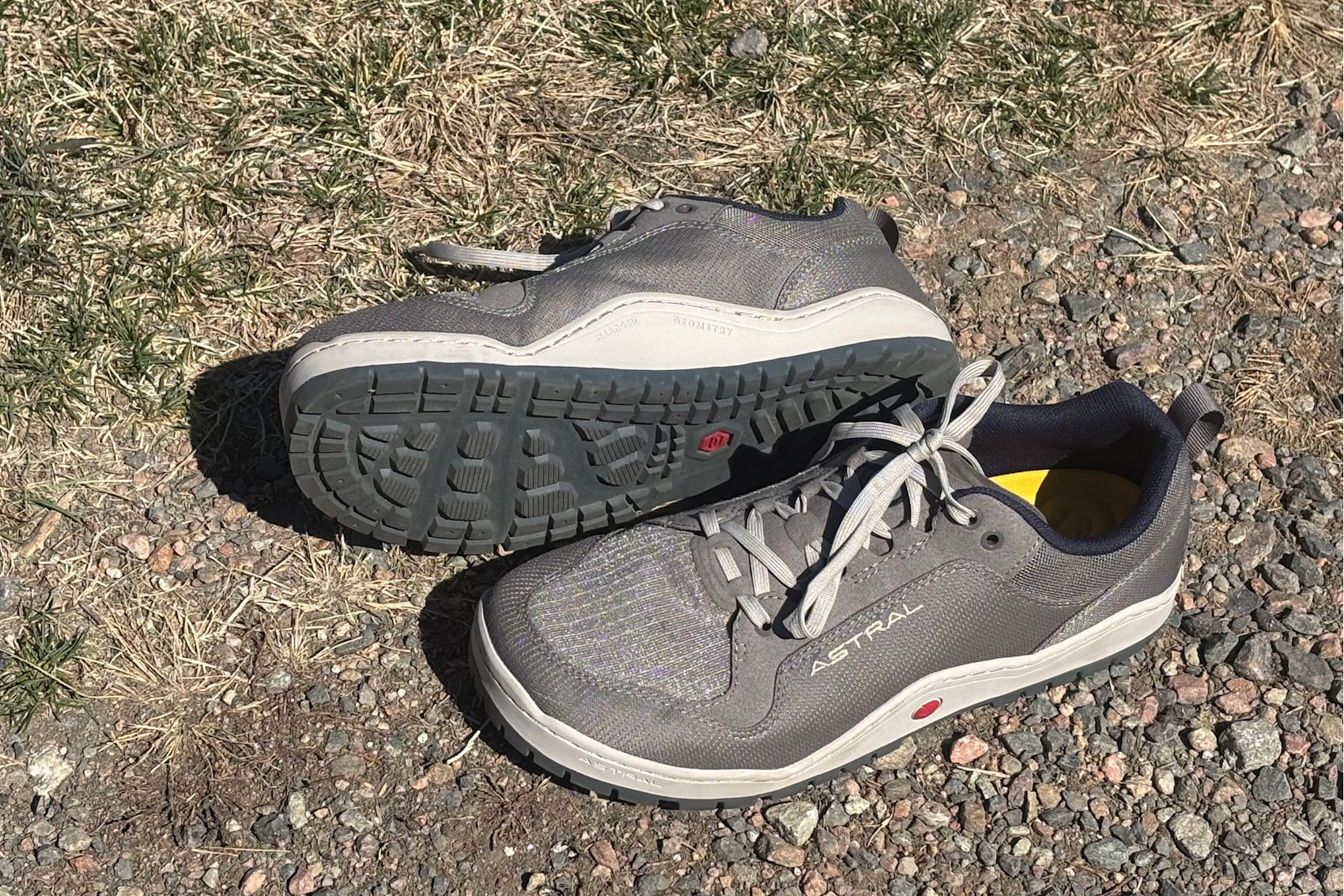 A pair of gray Astral shoes on a rocky, grassy surface in sunlight