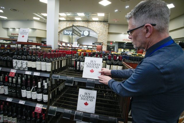 Canada-is-out-of-this-world-angry-at-Trumps-tariffs.jpg A grey haired man puts a sign with a red maple leaf on empty liquor store shelves that reads “Buy Canadian instead.”