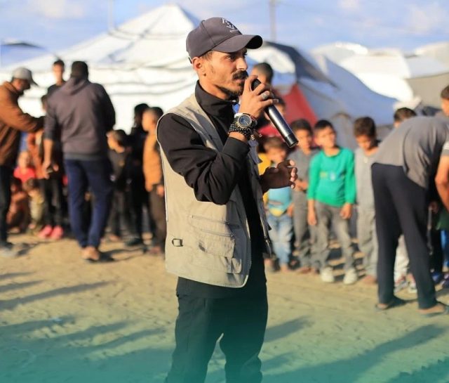 Palestinian-Artist-Dorgham-Quraiqi-Killed-in-Israeli-Airstrike.jpeg A man speaking into a microphone before a row of children.