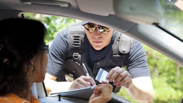 Study-of-Lyft-rideshare-data-confirms-minorities-get-more-tickets.jpg Image of a policeman leaning into the driver's side window of a car, writing a citation.