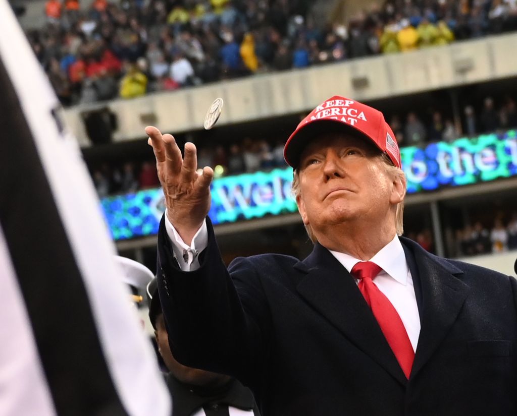 Trump-supporters-lose-billion-as-TRUMPs-value-falls-of-a.jpg A man in a suit and a red hat tosses a coin at a stadium.