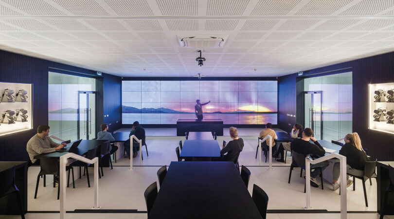 A modern classroom designed by WindsorPatania Architects features students seated at tables, a large video wall displaying a stunning sunset image, and a presenter standing in front.