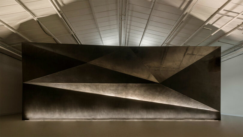 A large, dark metallic geometric wall display designed by WindsorPatania Architects features angular shapes lit from below, set in an industrial-style gallery space with a high ceiling.