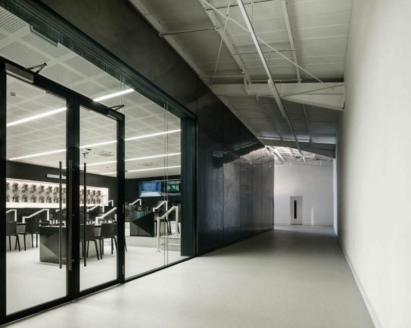 This modern corridor, crafted by WindsorPatania Architects, boasts glass doors on the left leading to a sleek, black-walled room with a counter and stools. The ceiling showcases exposed beams and pipes, embodying contemporary flair.