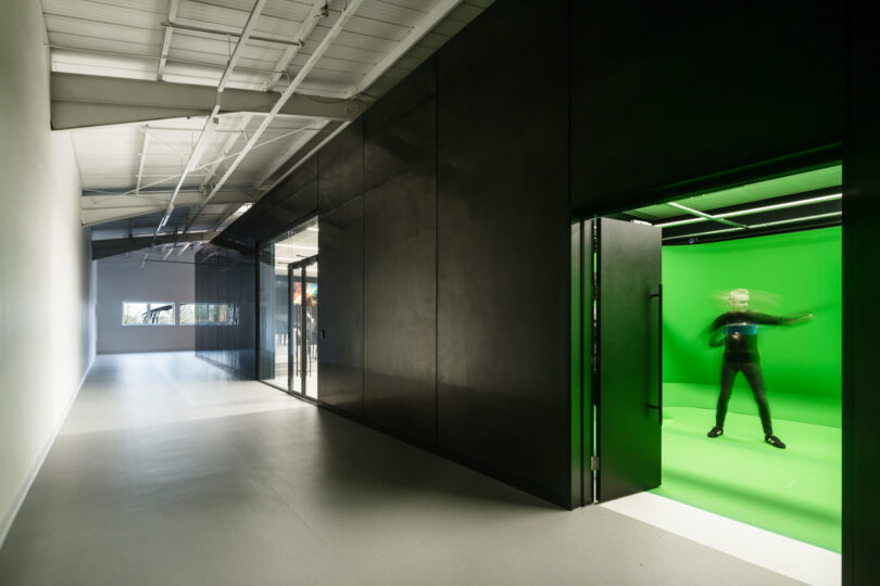 A person stands inside a green screen room with blurred motion. The room is part of a modern building interior with black walls and exposed ceiling structures.
