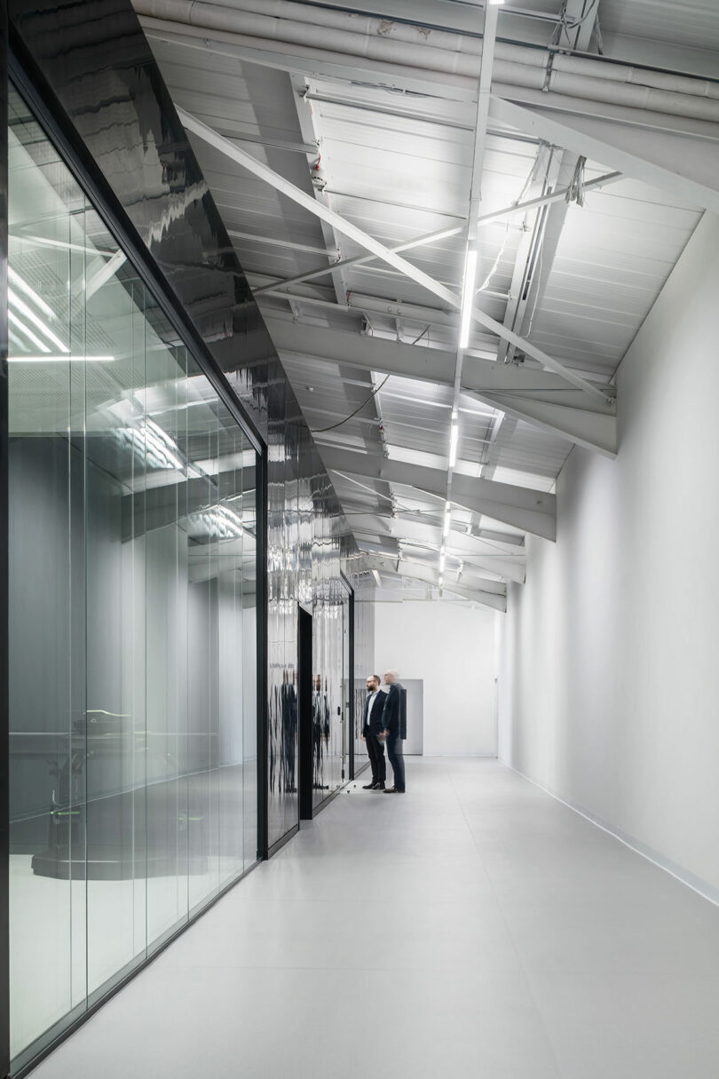 Three people stand together in a modern hallway designed by WindsorPatania Architects, with glass walls reflecting their presence. The ceiling boasts visible beams and lights, embodying a minimalistic and industrial vibe.