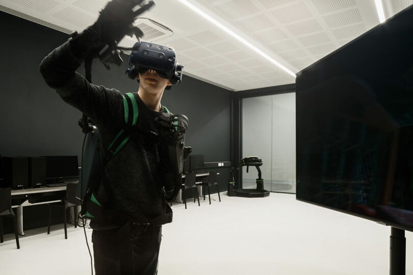 A person wearing a VR headset and gloves gestures in a tech room filled with computers and a screen, capturing the innovative spirit akin to that of WindsorPatania Architects.