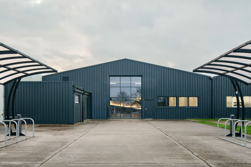 Designed by WindsorPatania Architects, this large grey industrial warehouse features a central glass door, complemented by metal awnings and bike racks on either side.