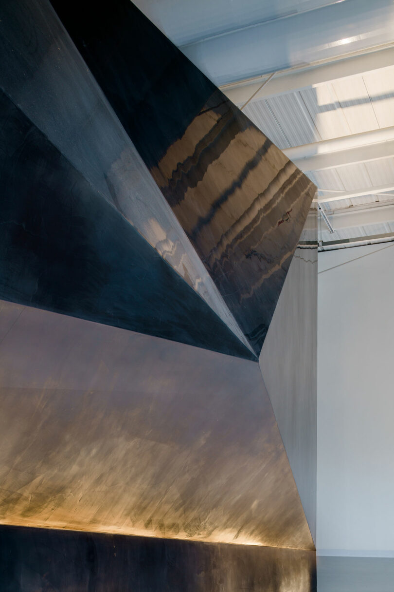 Sleek, angular metallic wall structure in an industrial space, designed by WindsorPatania Architects, lit from below. The area boasts a white ceiling adorned with beams and skylights.