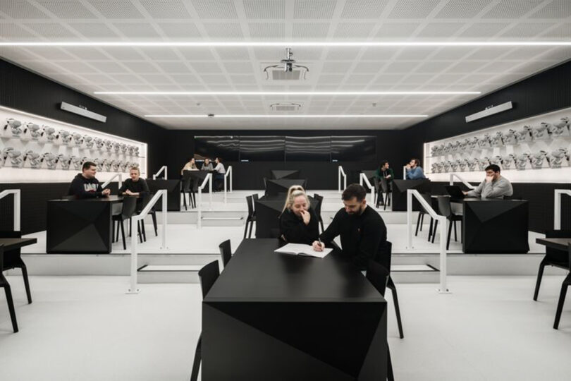 Modern classroom with students seated at black tables, surrounded by white walls displaying animal skull models. The room features geometric design elements and bright lighting.