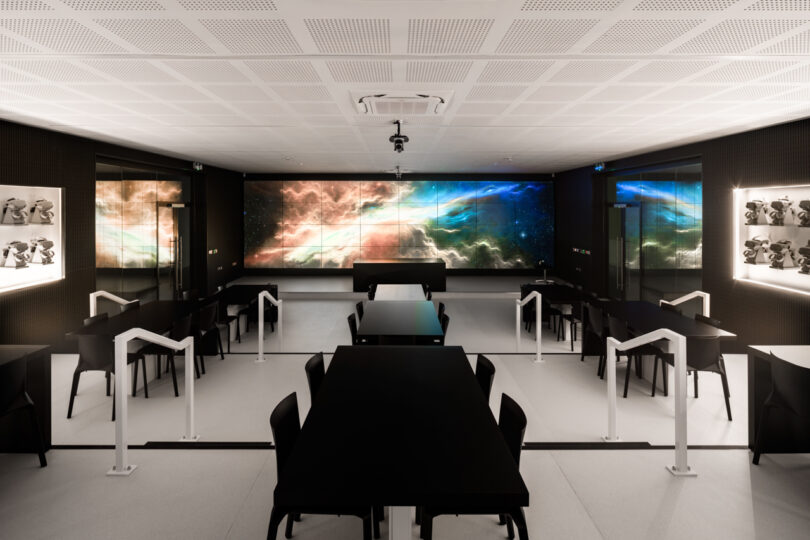 A modern room with multiple black tables and chairs, large screens displaying colorful nebula images on the walls, and white barriers separating sections.