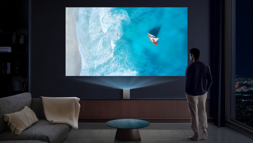 A person stands in a dimly lit room watching a large screen displaying an aerial view of a beach with a small boat on blue water.