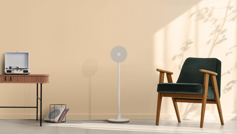 Minimalist living room with a chair, a standing fan, and a table with a record player and magazine rack. Shadows of leaves are cast on the beige wall.