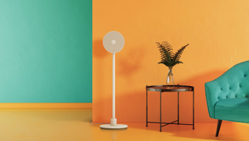 A standing fan on a yellow floor next to a small round table with a plant, set against orange and green walls and a turquoise sofa.