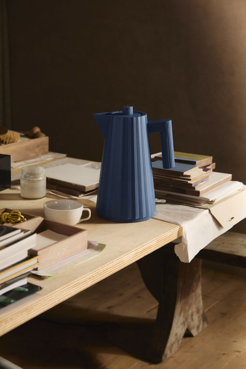 A blue coffee carafe sits on a wooden table surrounded by notebooks, a white cup, and other desk items