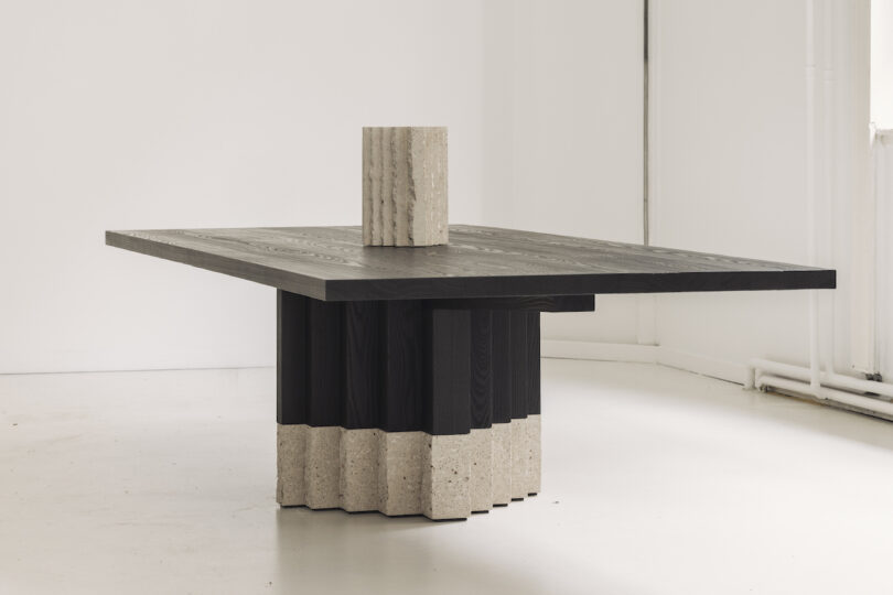 A modern square table with a black wooden top and a textured stone cylindrical base in a minimalist white room