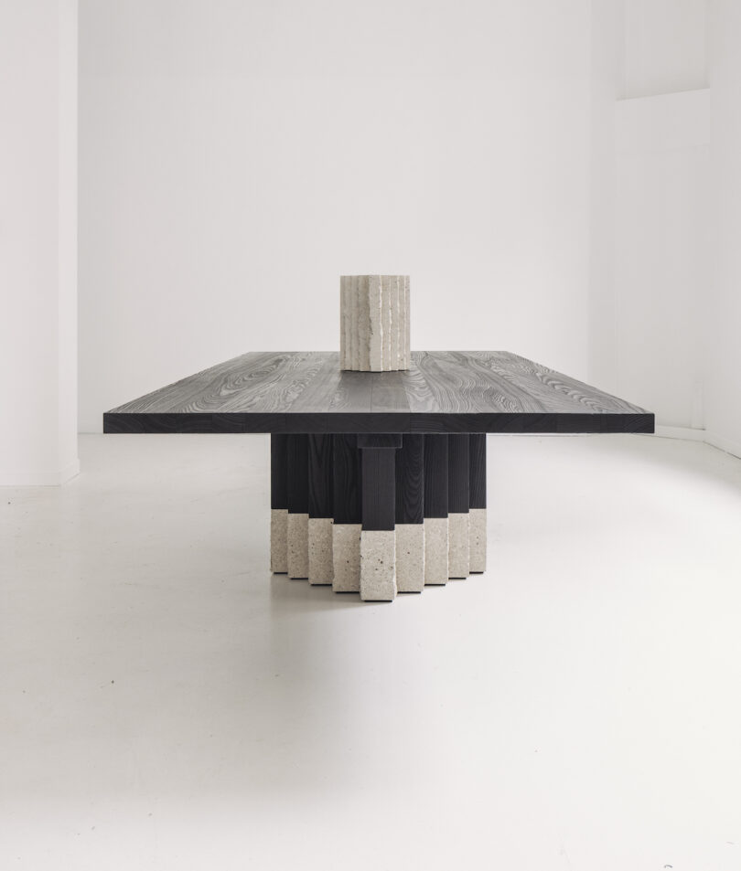 A modern square table with a black wooden top and a textured stone cylindrical base in a minimalist white room