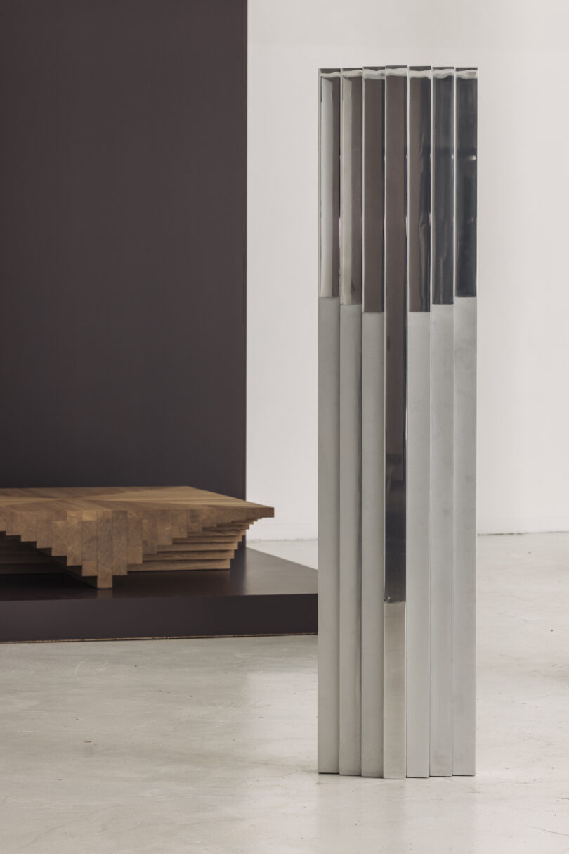A vertical metal sculpture with parallel rectangular panels stands on a concrete floor. A brown, tiered wooden structure is partially visible in the background