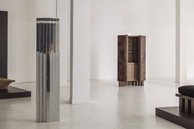 A minimalist gallery space with a silver, vertical art piece on the left, wooden furniture on dark platforms, and a patterned cabinet against a white wall