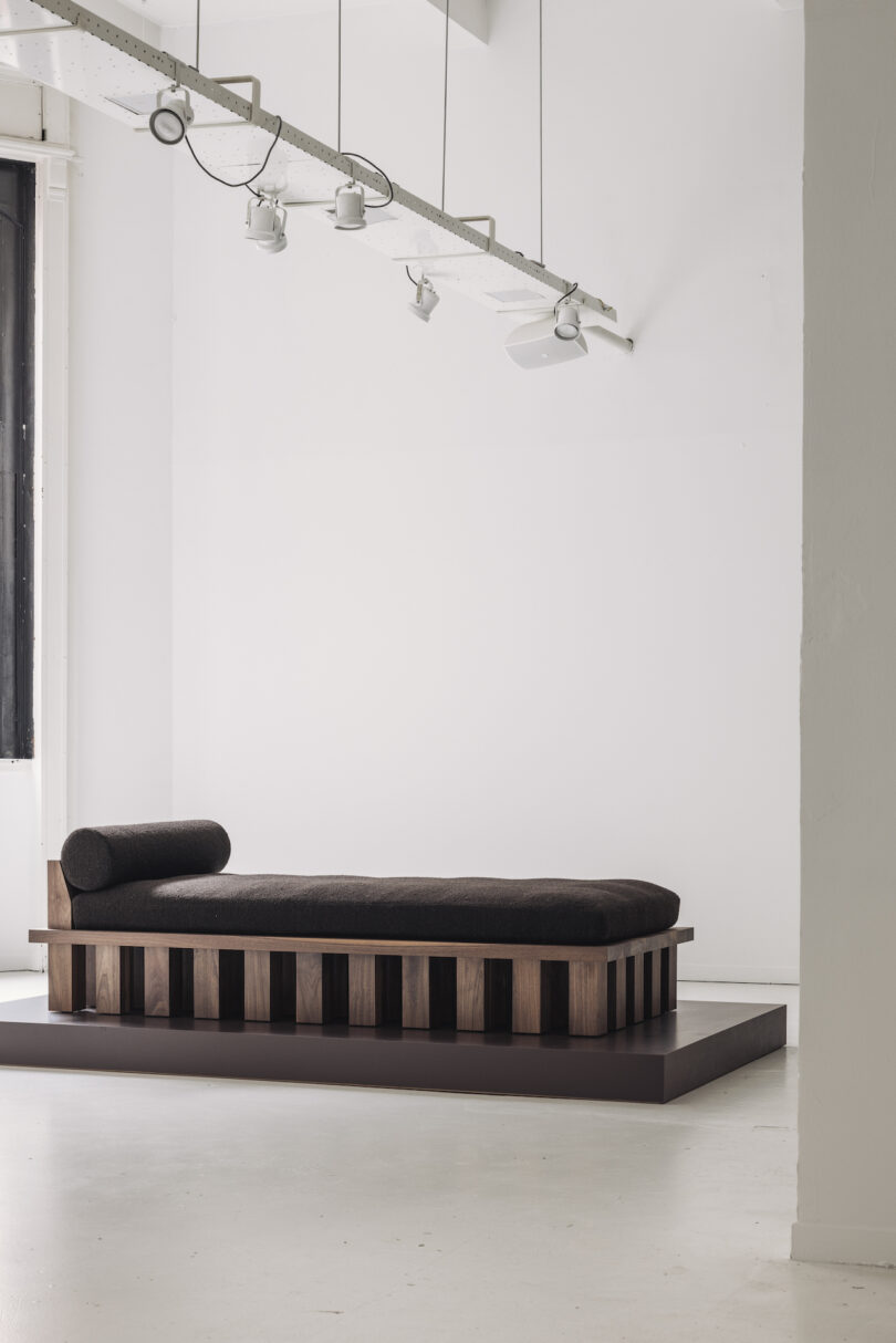 Modern daybed with dark cushion and wooden slatted base on a raised platform in a minimal, white room with ceiling spotlights