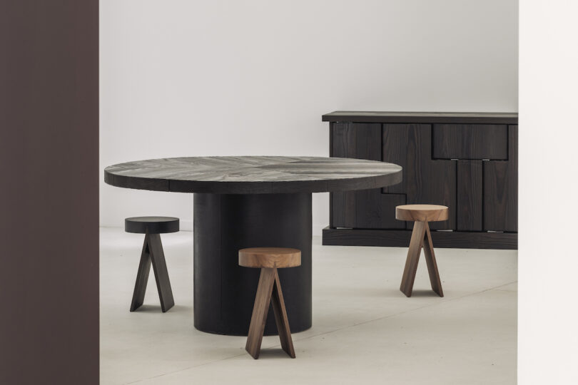 A minimalist room with a round black table, three wooden stools, and a dark sideboard in the background