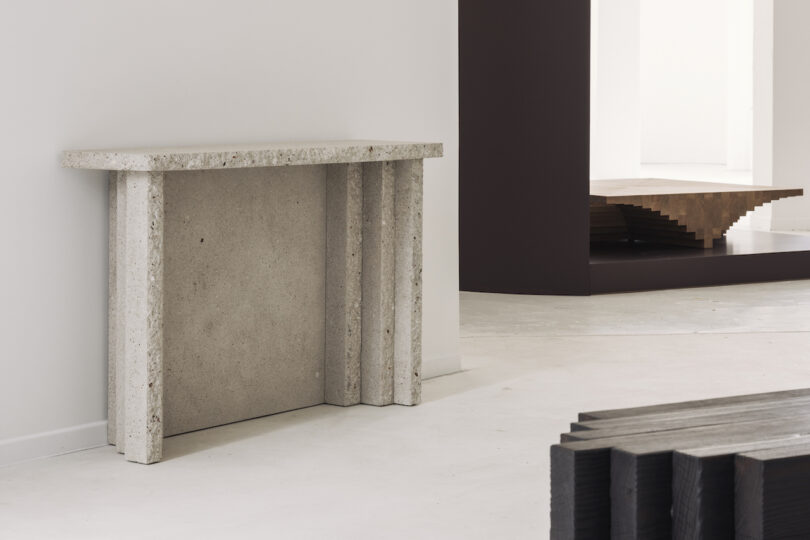 Minimalist stone console table against a white wall, next to an abstract wooden staircase and a black bench in a modern gallery space
