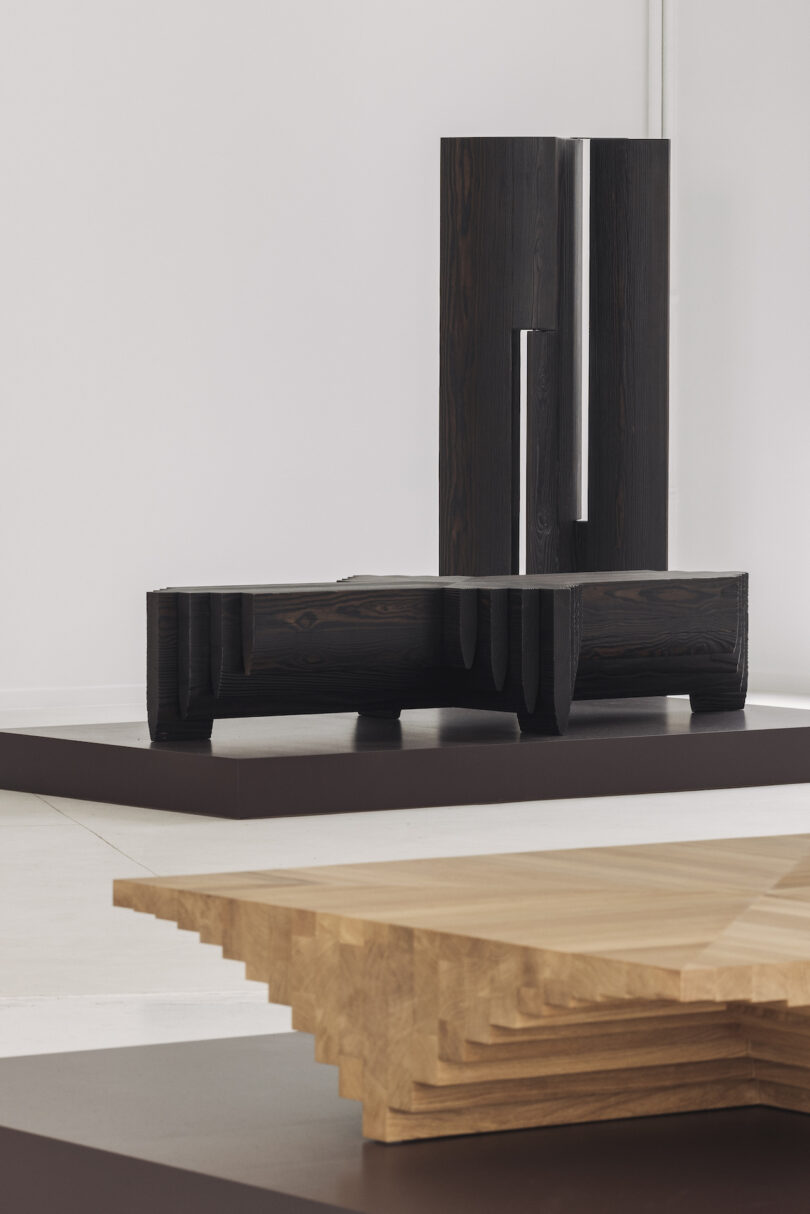 Geometric wooden sculptures displayed in a minimalist, white gallery space. One sculpture is dark and vertical; the other is light and horizontal with stepped layers