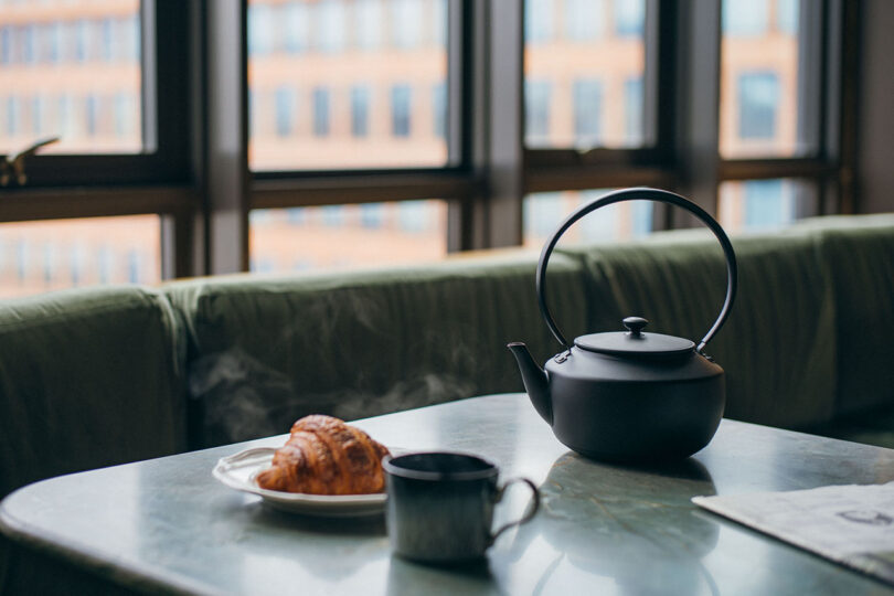Black teapot, cup, and croissant on a table near a window with blurred buildings in the background.