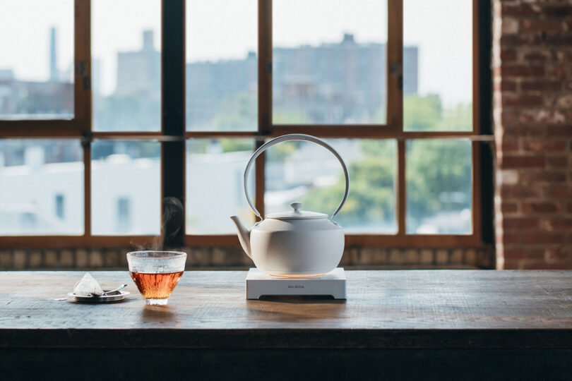 A white teapot on a warmer and a glass cup of tea on a saucer are on a wooden table by large windows with an urban view.