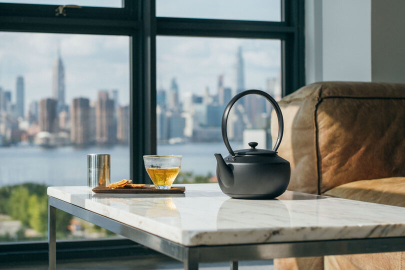 A black teapot, glass cup, and cookies on a tray are on a marble table by a large window, overlooking a city skyline.