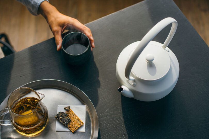 A white teapot and a tray with a clear pitcher of tea, crackers, and a hand holding a dark cup on a dark table.