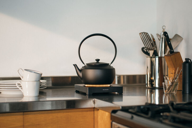 Black kettle on an induction stove in a kitchen with metal countertop, white plates, stacked white mugs, and utensils in a holder.