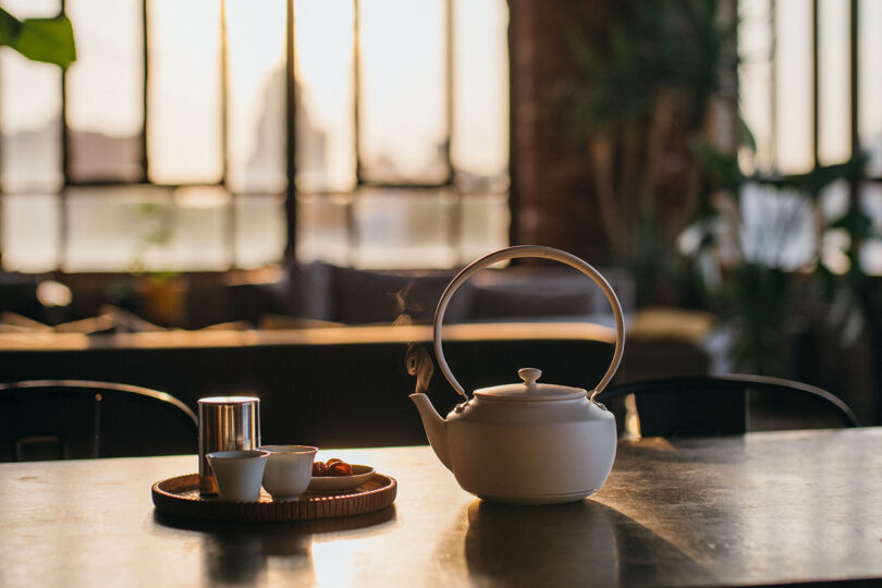 A white teapot on a table beside a tray with two cups and a small container. Sunlight streams through large windows in the background, creating a warm ambiance.