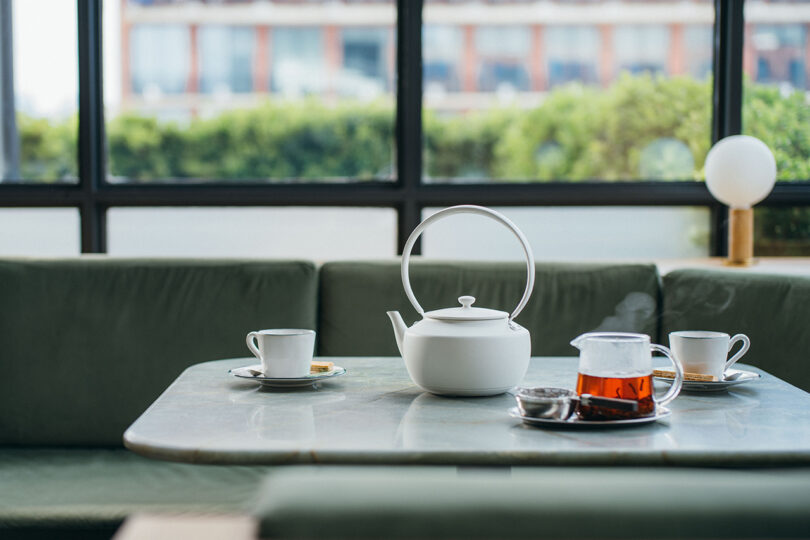 White teapot and clear glass of tea on a table with two cups and saucers in a modern setting with large windows.
