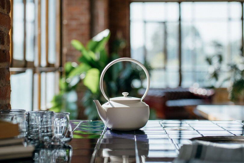 White teapot with a large handle on a sunlit tiled counter, surrounded by glass cups and green plants in the background.