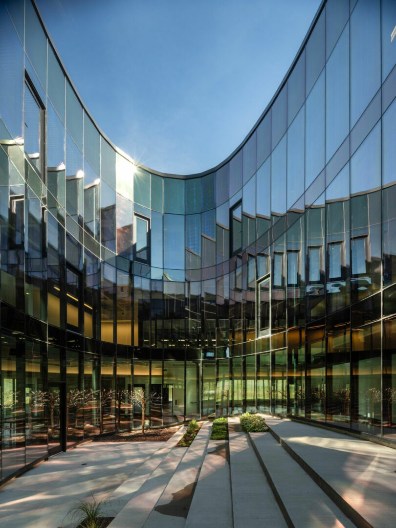 Designed by CLB Architects, this modern glass building features a stunning curved facade that masterfully reflects the sky. The exterior view showcases expansive windows and a few small plants lining a paved path in the foreground.