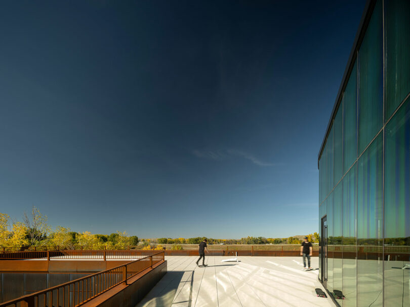 Two people stand on a rooftop terrace, the hallmark design of CLB Architects, with a stunning glass building and lush trees in the background under a clear blue sky.