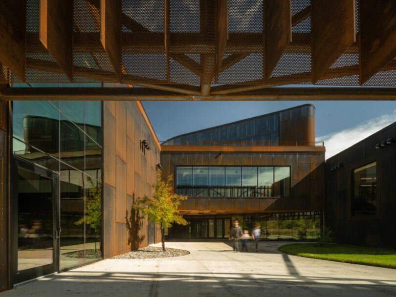 Designed by CLB Architects, the modern building boasts a rust-colored metal exterior and large glass windows. A landscaped courtyard invites visitors, while two blurred figures stroll along a concrete pathway under a detailed metal overhang.