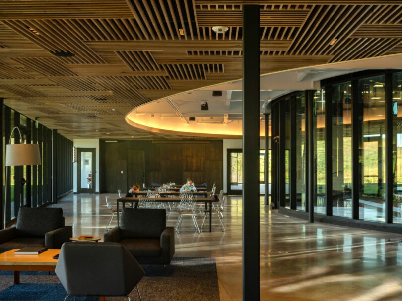 The modern office space, designed by CLB Architects, features a wooden ceiling, large windows, lounge chairs, and a communal table. Two people sit at the table in this spacious, well-lit environment.