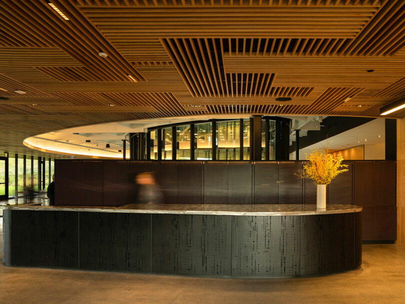 The modern lobby, designed by CLB Architects, features a curved, dark reception desk with a marble top. A vase with yellow flowers adds a splash of color. Wooden ceiling panels and glass walls set the background as a person walks by.